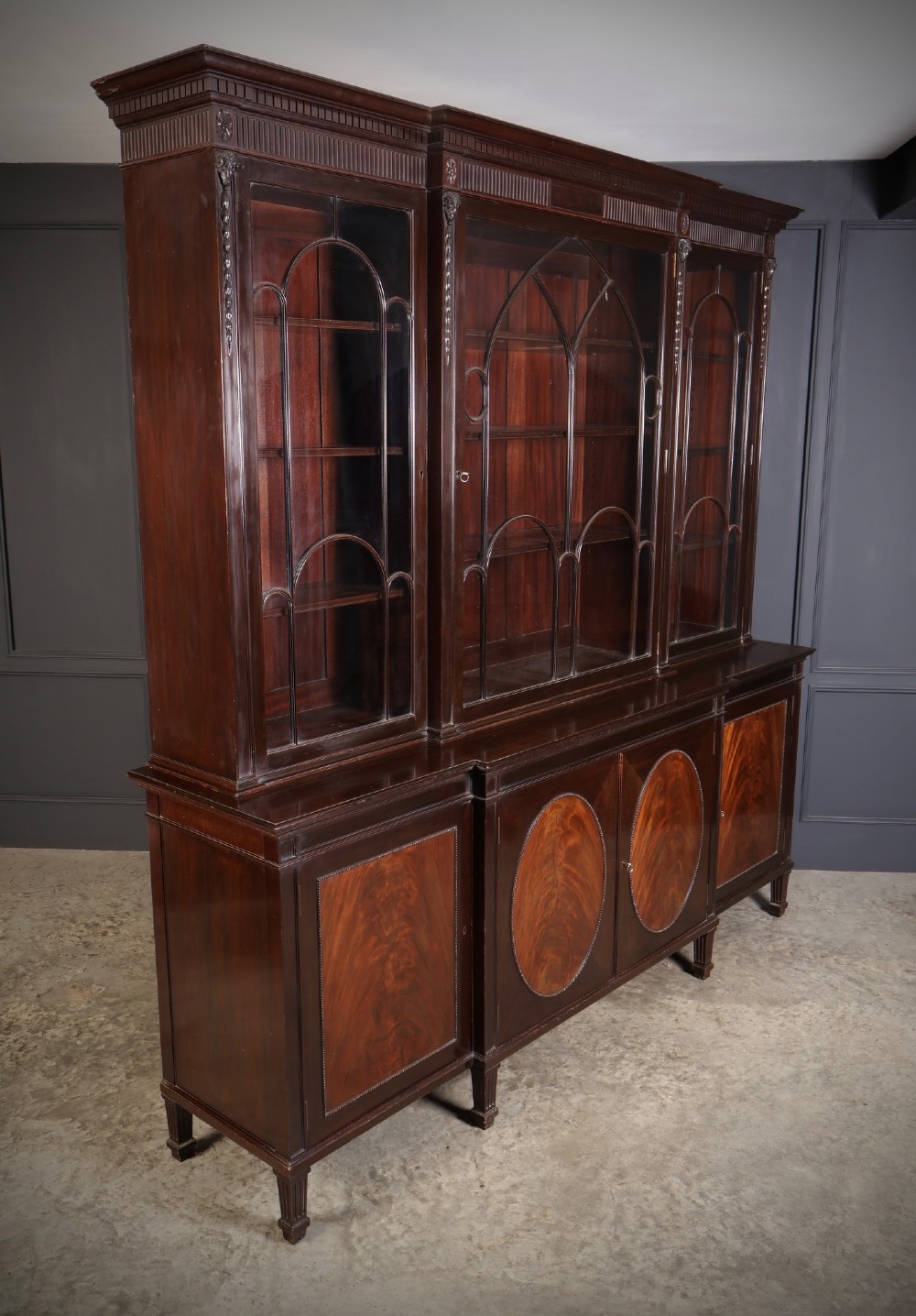 A large mahogany breakfront bookcase featuring upper glazed doors with astragal tracery above lower cupboards with oval flame mahogany veneer panels. View II