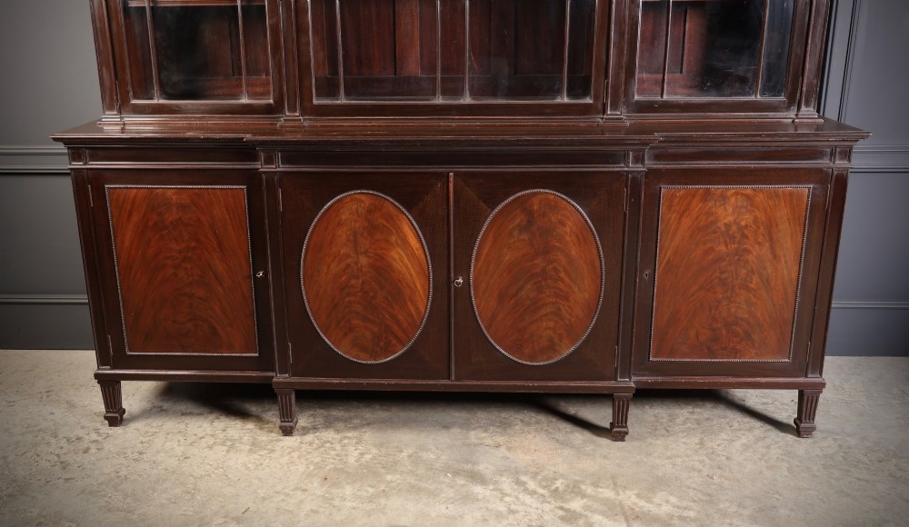 A large mahogany breakfront bookcase featuring upper glazed doors with astragal tracery above lower cupboards with oval flame mahogany veneer panels. View IV