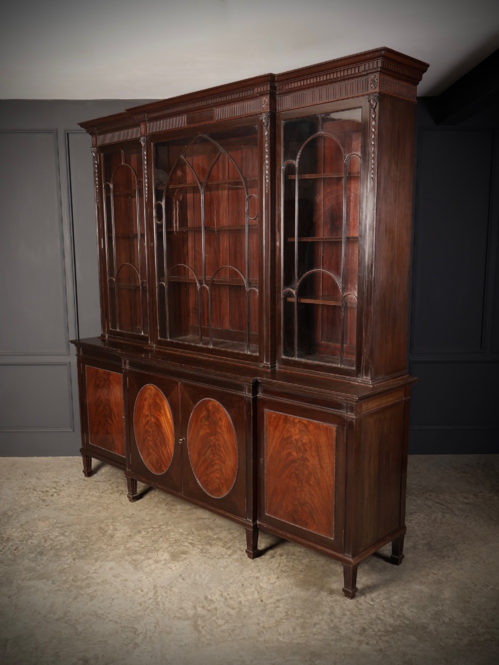 A large mahogany breakfront bookcase featuring upper glazed doors with astragal tracery above lower cupboards with oval flame mahogany veneer panels. View VI
