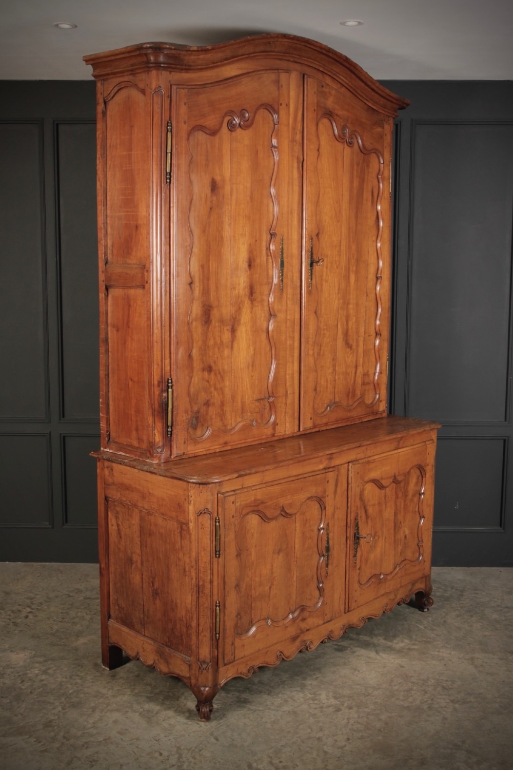 An 18th-century French Provincial two-tier cherry wood cupboard featuring moulded panel doors, long brass hinges, and a curved top cornice.