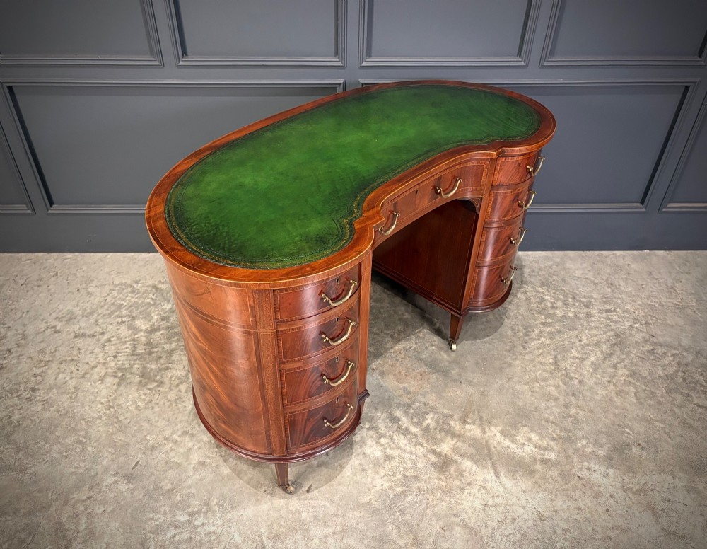 An Edwardian mahogany kidney-shaped writing desk with an inset green leather top, satinwood inlay, curved drawers, brass handles, and tapered legs on castors.