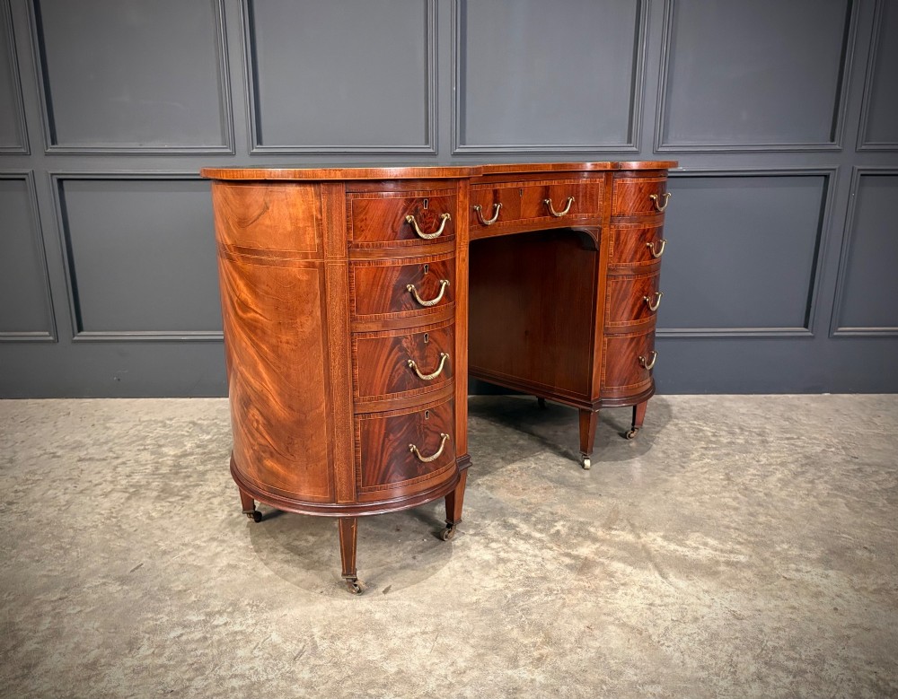 An Edwardian mahogany kidney-shaped writing desk with an inset green leather top, satinwood inlay, curved drawers, brass handles, and tapered legs on castors. View II