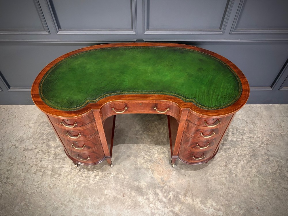 An Edwardian mahogany kidney-shaped writing desk with an inset green leather top, satinwood inlay, curved drawers, brass handles, and tapered legs on castors. View V