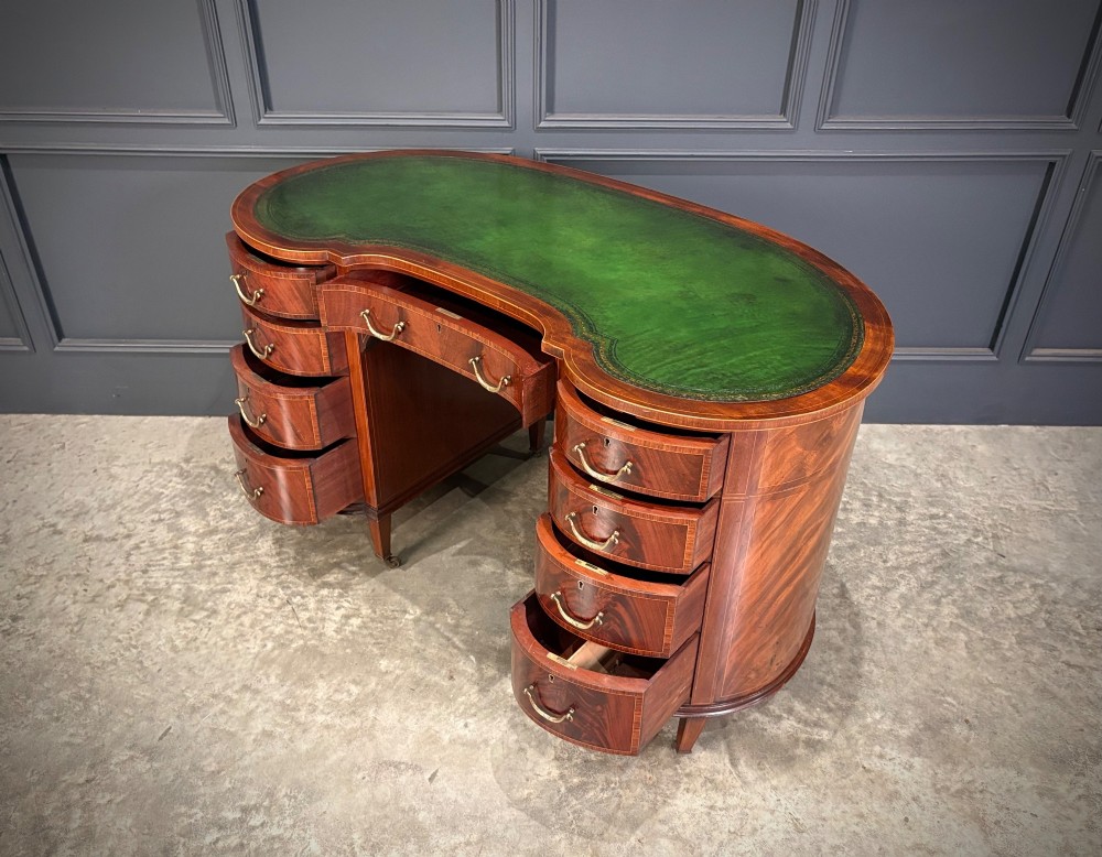An Edwardian mahogany kidney-shaped writing desk with an inset green leather top, satinwood inlay, curved drawers, brass handles, and tapered legs on castors. View VI