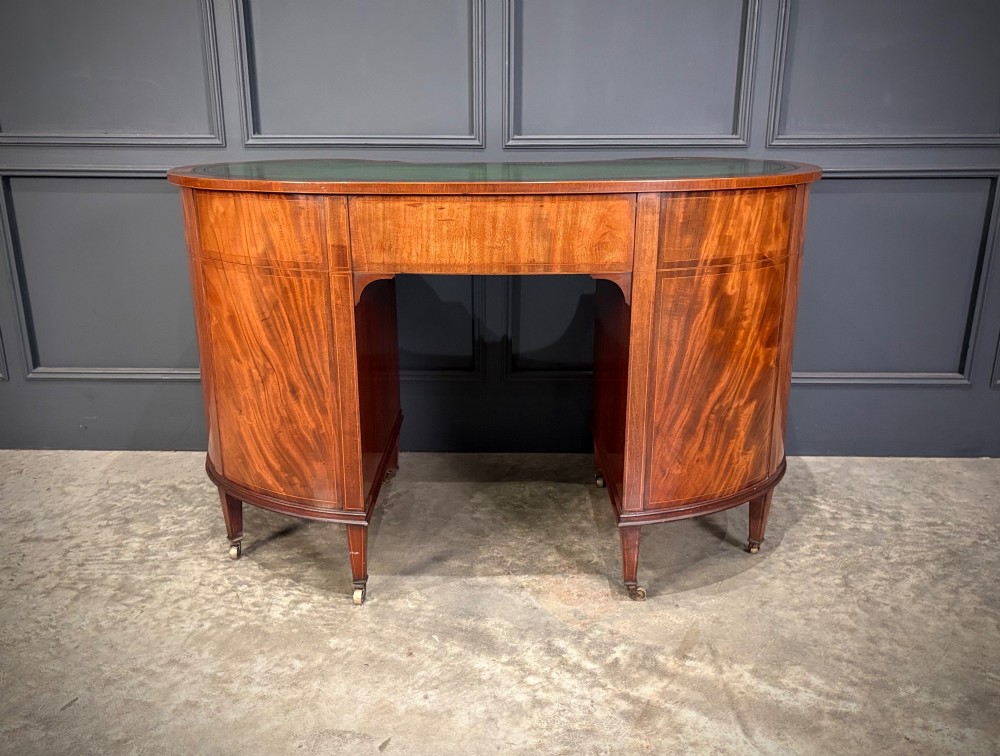 An Edwardian mahogany kidney-shaped writing desk with an inset green leather top, satinwood inlay, curved drawers, brass handles, and tapered legs on castors. View VIII