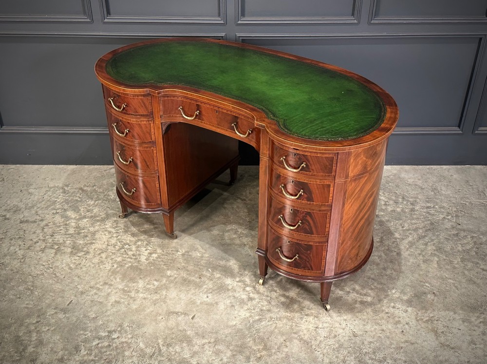 An Edwardian mahogany kidney-shaped writing desk with an inset green leather top, satinwood inlay, curved drawers, brass handles, and tapered legs on castors. View XI