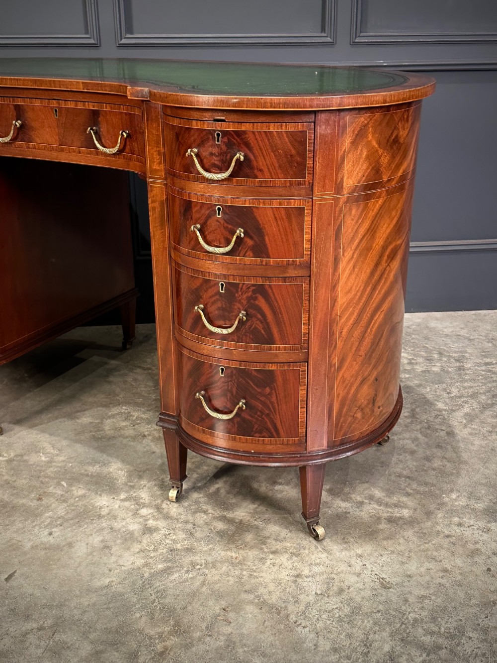 An Edwardian mahogany kidney-shaped writing desk with an inset green leather top, satinwood inlay, curved drawers, brass handles, and tapered legs on castors. View XIII