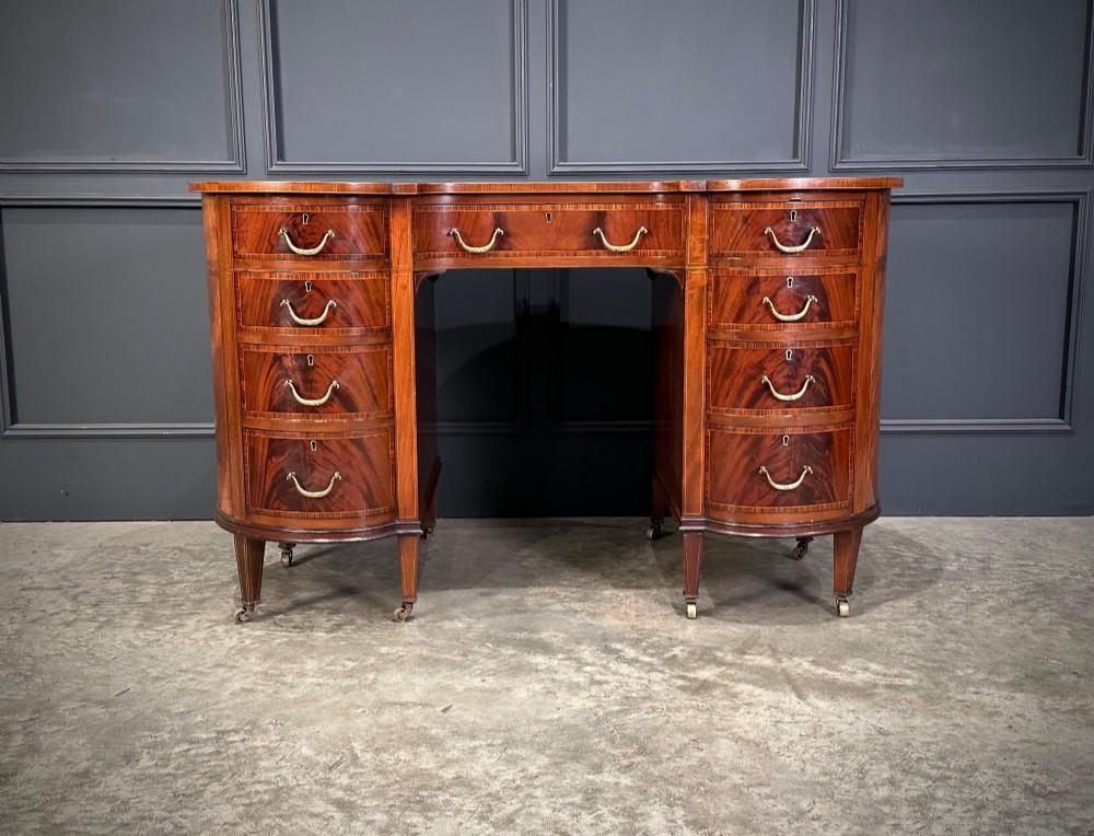 An Edwardian mahogany kidney-shaped writing desk with an inset green leather top, satinwood inlay, curved drawers, brass handles, and tapered legs on castors. View XIV