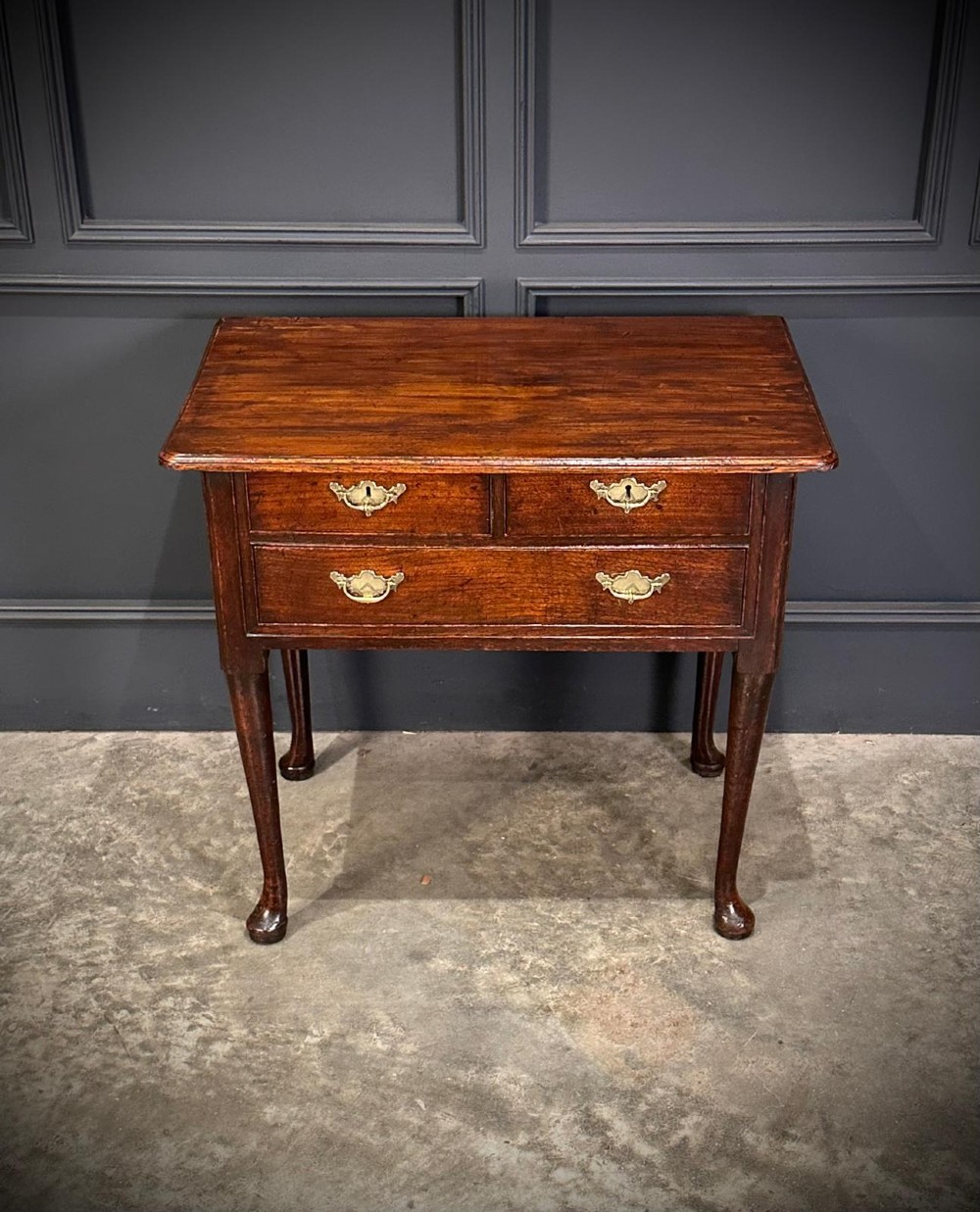 A Queen Anne dark oak lowboy featuring three drawers with brass backplate handles resting on slender cabriole legs with pad feet.
