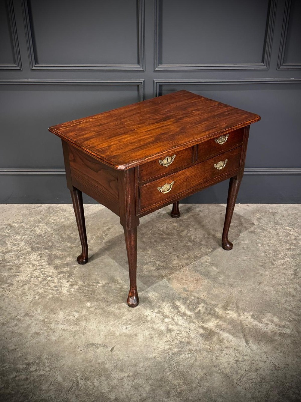 A Queen Anne dark oak lowboy featuring three drawers with brass backplate handles resting on slender cabriole legs with pad feet. View IV