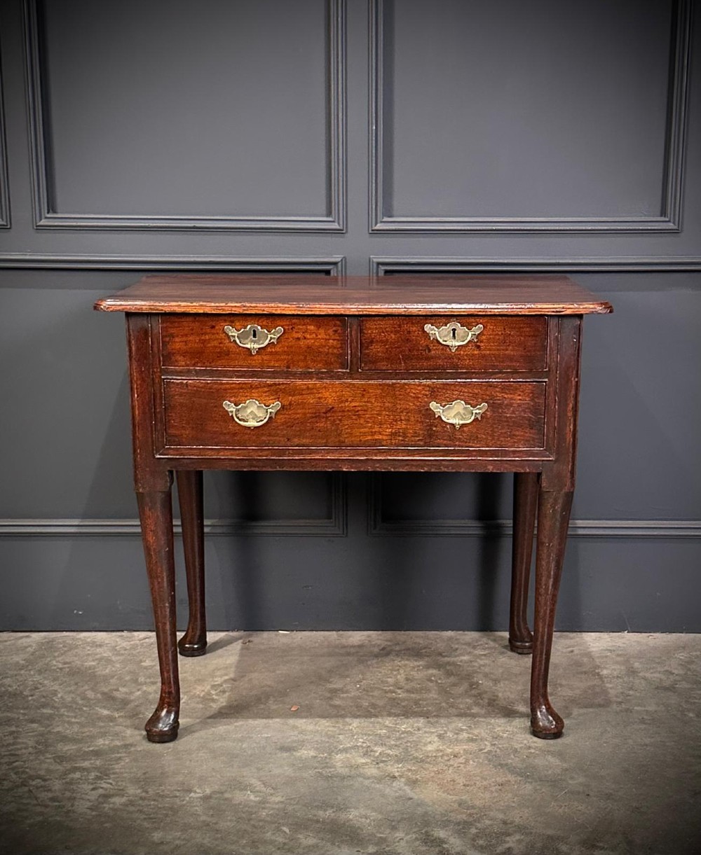 A Queen Anne dark oak lowboy featuring three drawers with brass backplate handles resting on slender cabriole legs with pad feet. View V