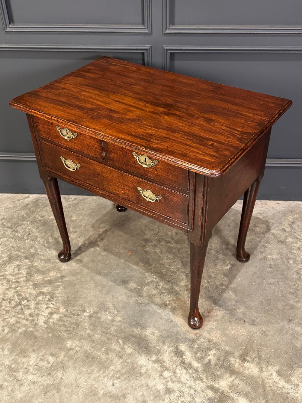 A Queen Anne dark oak lowboy featuring three drawers with brass backplate handles resting on slender cabriole legs with pad feet. View VI