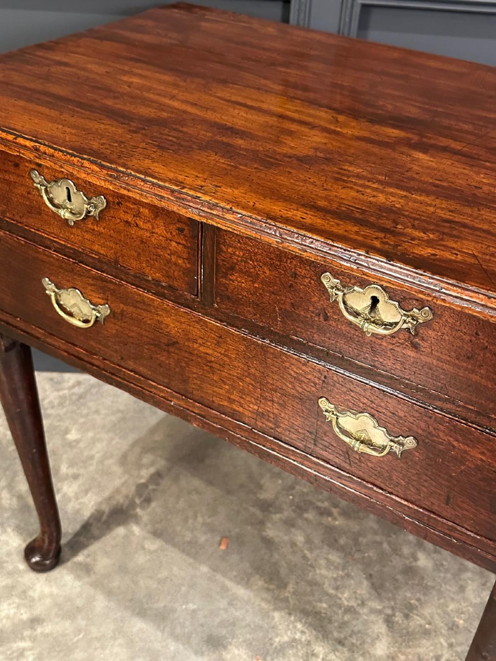 A Queen Anne dark oak lowboy featuring three drawers with brass backplate handles resting on slender cabriole legs with pad feet. View VII