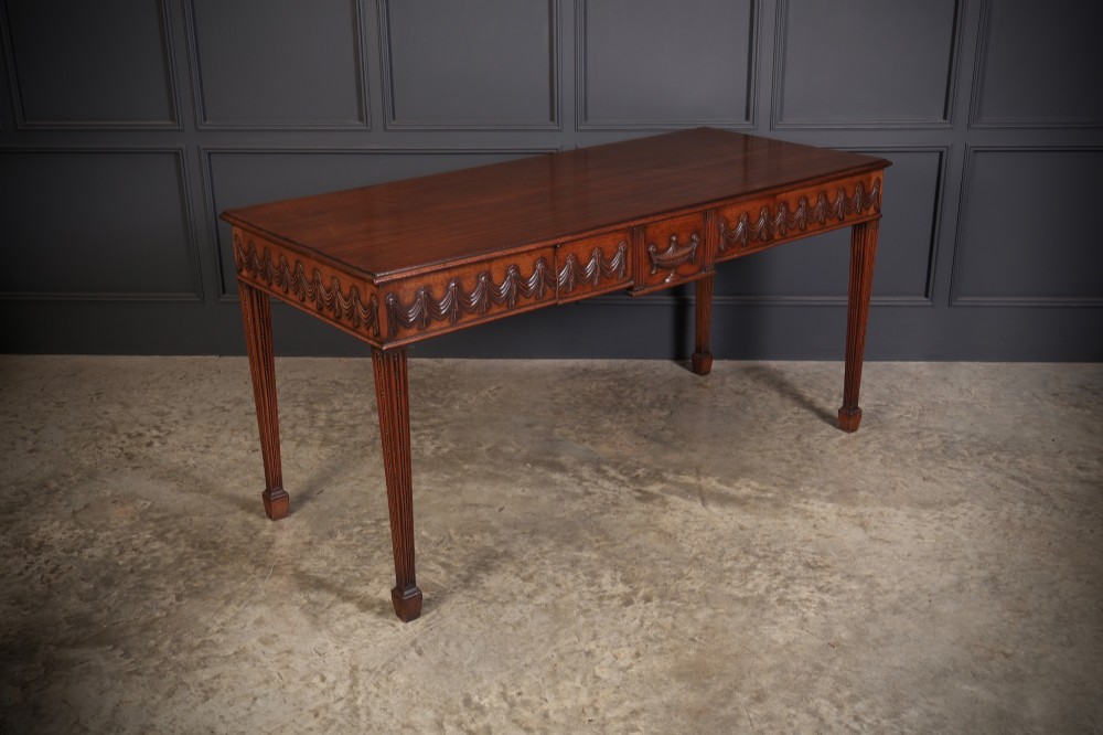 A large Sheraton style mahogany serving table featuring a rectangular top, frieze carved with drapery swags and a central urn relief, supported by four tapered fluted legs. View II
