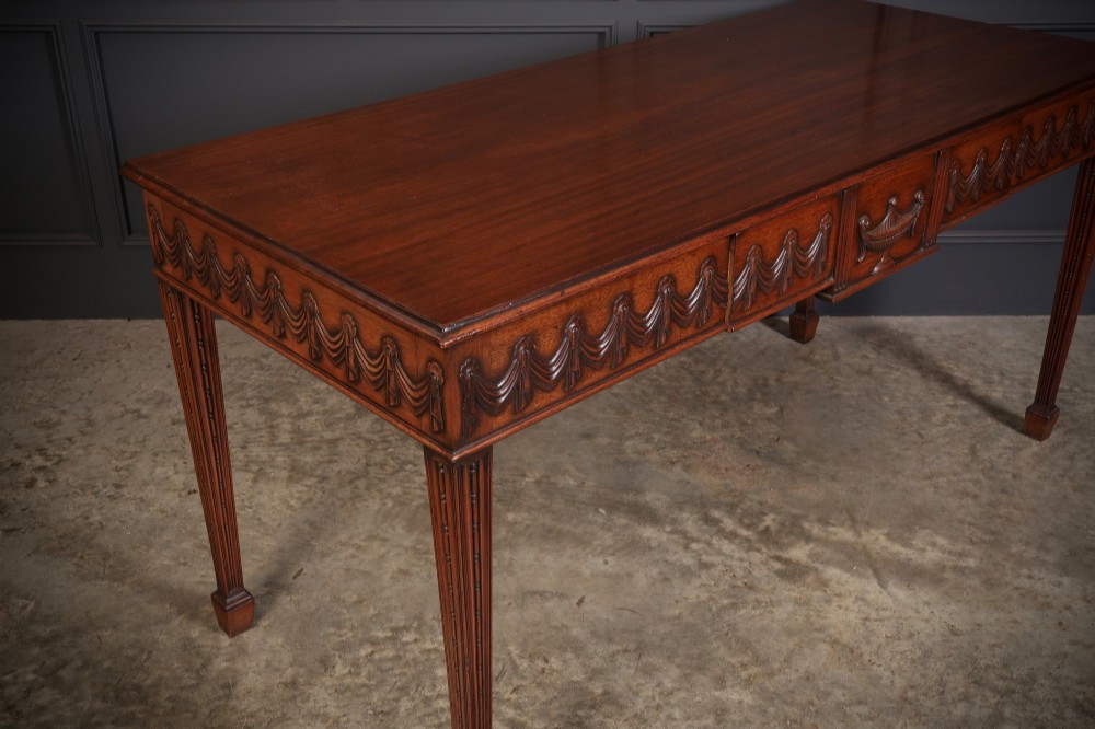 A large Sheraton style mahogany serving table featuring a rectangular top, frieze carved with drapery swags and a central urn relief, supported by four tapered fluted legs. View III