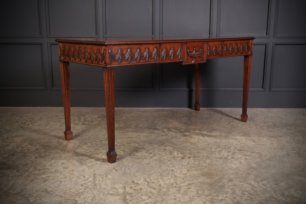 A large Sheraton style mahogany serving table featuring a rectangular top, frieze carved with drapery swags and a central urn relief, supported by four tapered fluted legs. View IV