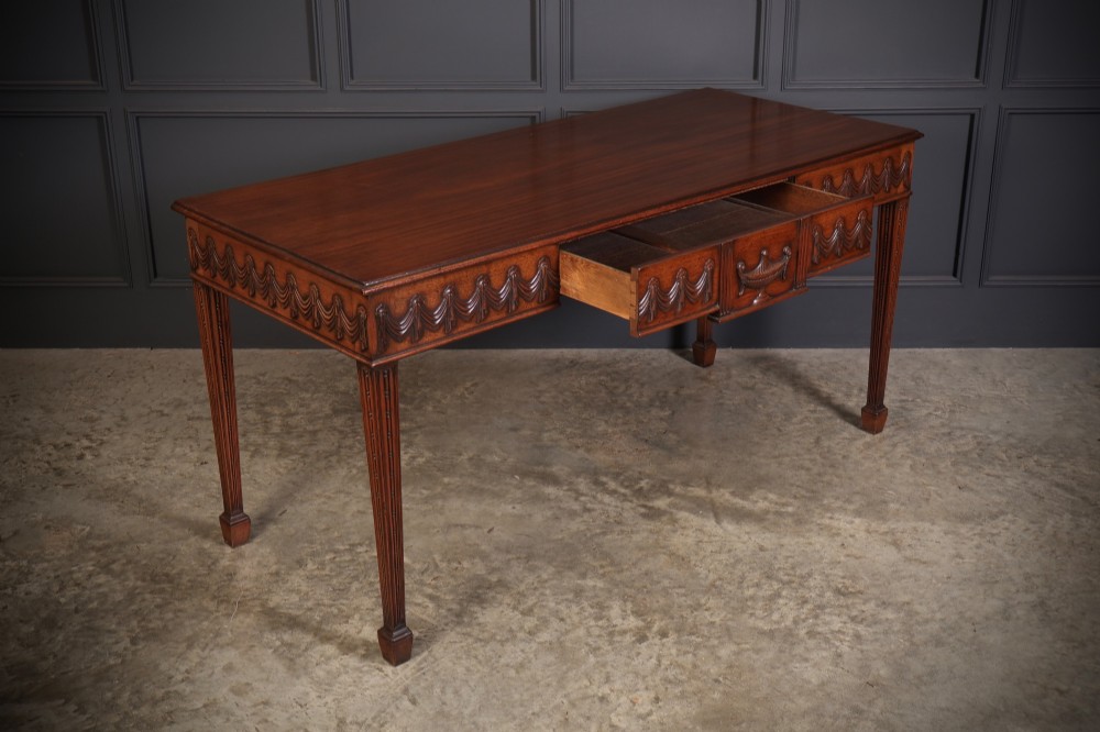 A large Sheraton style mahogany serving table featuring a rectangular top, frieze carved with drapery swags and a central urn relief, supported by four tapered fluted legs. View V