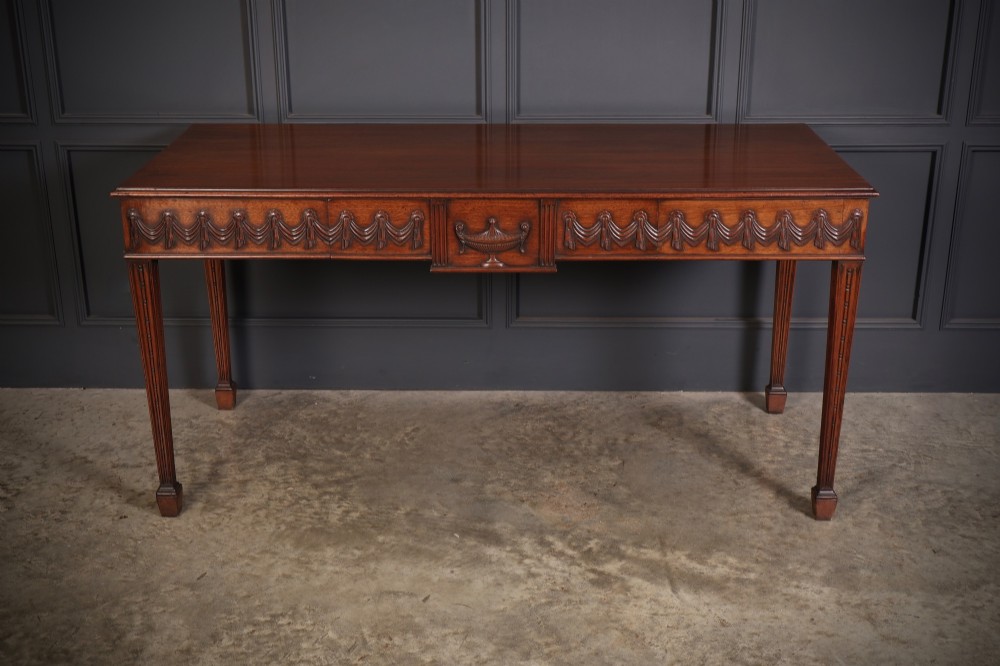 A large Sheraton style mahogany serving table featuring a rectangular top, frieze carved with drapery swags and a central urn relief, supported by four tapered fluted legs.