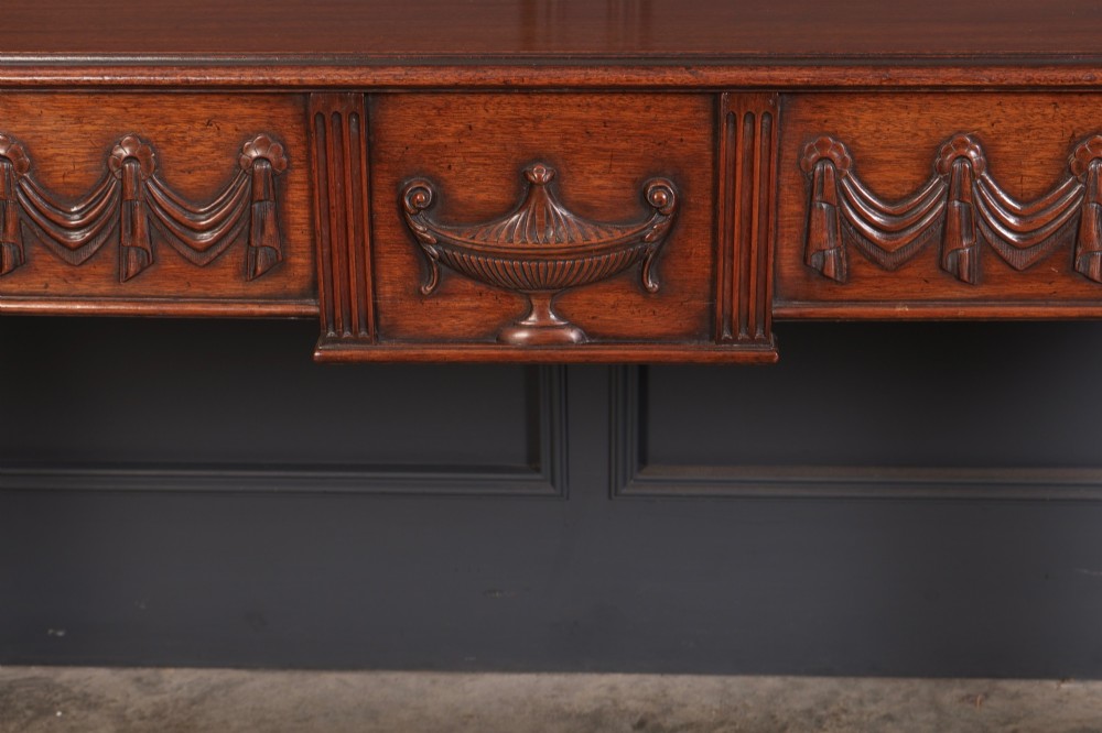 A large Sheraton style mahogany serving table featuring a rectangular top, frieze carved with drapery swags and a central urn relief, supported by four tapered fluted legs. View VI