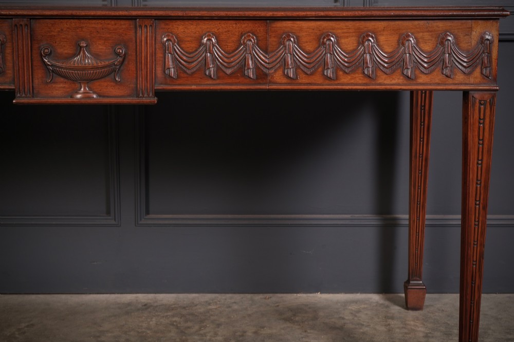 A large Sheraton style mahogany serving table featuring a rectangular top, frieze carved with drapery swags and a central urn relief, supported by four tapered fluted legs. View VII