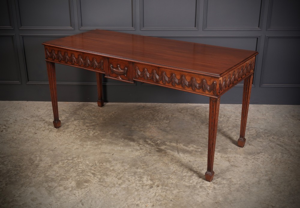 A large Sheraton style mahogany serving table featuring a rectangular top, frieze carved with drapery swags and a central urn relief, supported by four tapered fluted legs. View IX