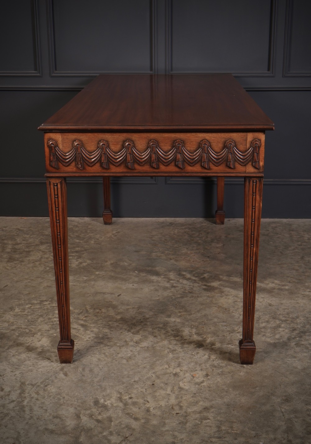 A large Sheraton style mahogany serving table featuring a rectangular top, frieze carved with drapery swags and a central urn relief, supported by four tapered fluted legs. View X