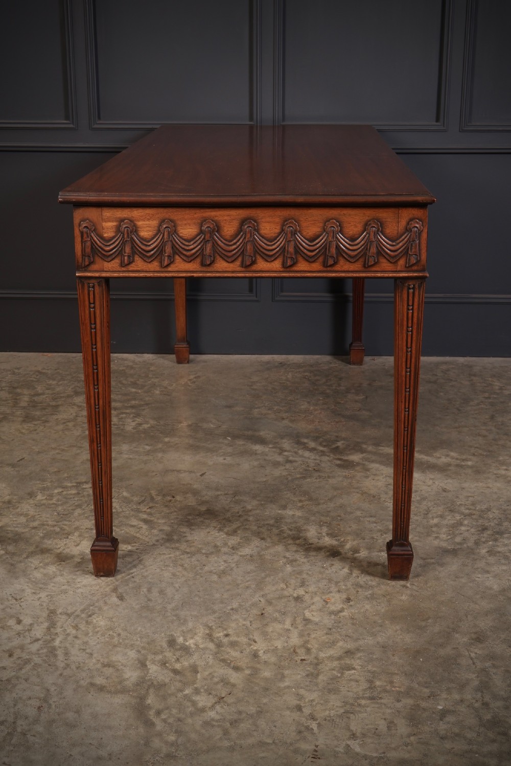 A large Sheraton style mahogany serving table featuring a rectangular top, frieze carved with drapery swags and a central urn relief, supported by four tapered fluted legs. View XII
