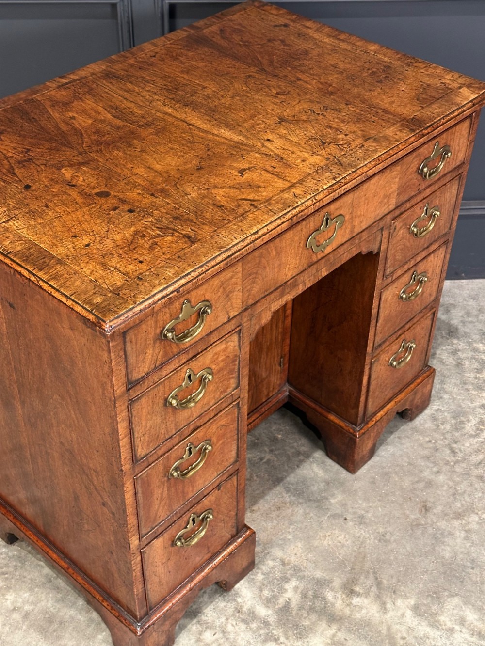 An 18th-century walnut kneehole desk featuring a flat top, multiple small drawers with brass drop handles, a recessed central cupboard, and bracket feet. View IV