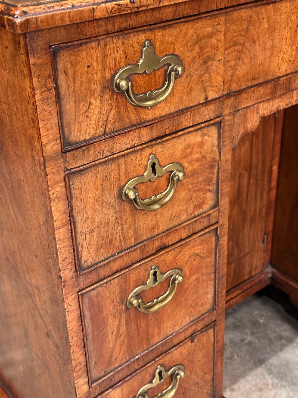 An 18th-century walnut kneehole desk featuring a flat top, multiple small drawers with brass drop handles, a recessed central cupboard, and bracket feet. View V