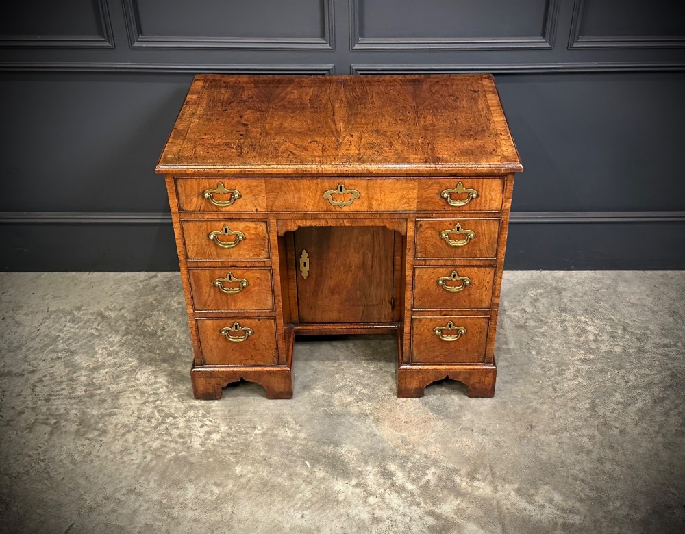 An 18th-century walnut kneehole desk featuring a flat top, multiple small drawers with brass drop handles, a recessed central cupboard, and bracket feet. View VI