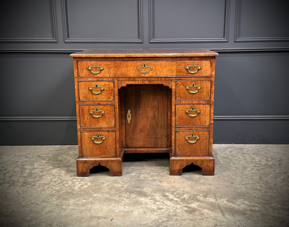 An 18th-century walnut kneehole desk featuring a flat top, multiple small drawers with brass drop handles, a recessed central cupboard, and bracket feet. View VII