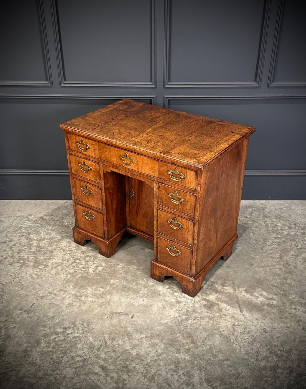 An 18th-century walnut kneehole desk featuring a flat top, multiple small drawers with brass drop handles, a recessed central cupboard, and bracket feet. View IX