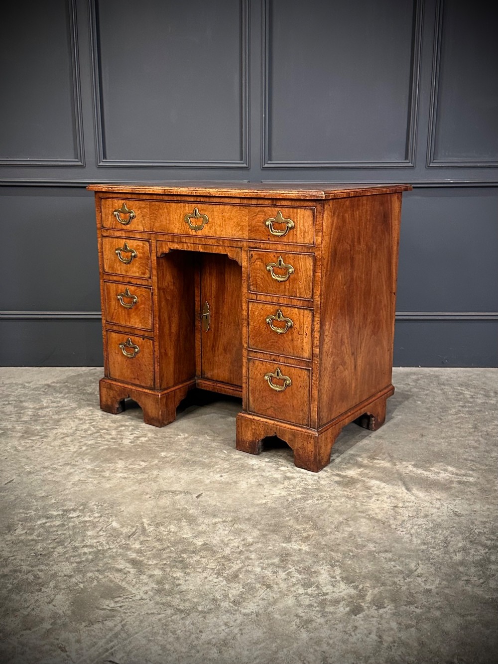 An 18th-century walnut kneehole desk featuring a flat top, multiple small drawers with brass drop handles, a recessed central cupboard, and bracket feet. View X