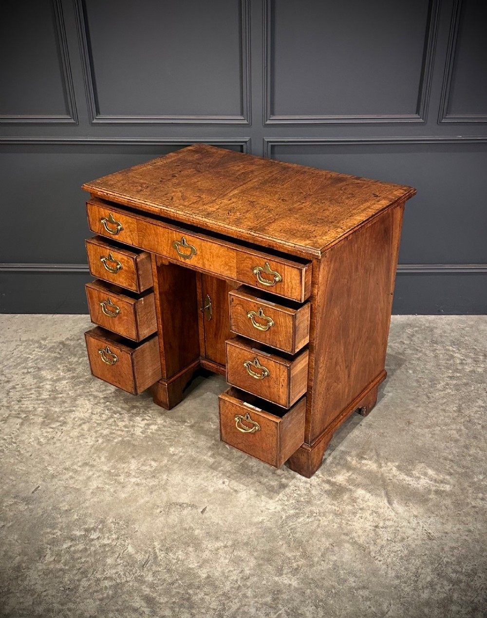 An 18th-century walnut kneehole desk featuring a flat top, multiple small drawers with brass drop handles, a recessed central cupboard, and bracket feet. View XI