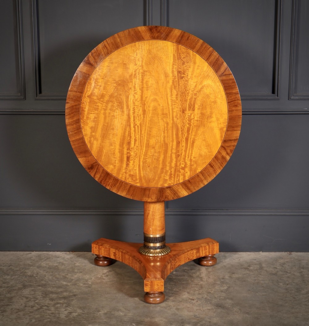 A circular antique satinwood occasional table featuring a crossbanded timber top, a central cylindrical pedestal with a brass mount, and a triform base on bun feet. View II