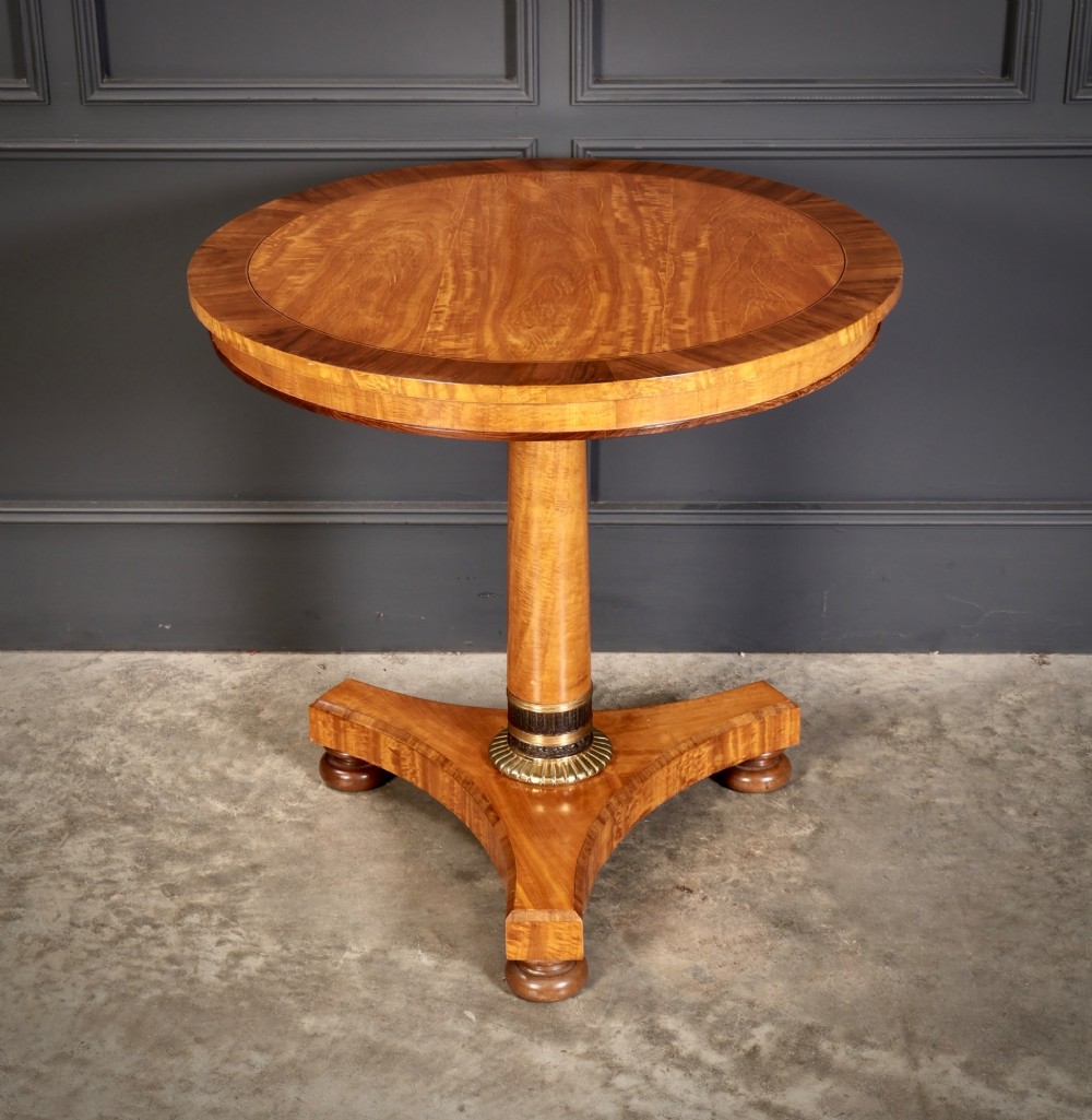 A circular antique satinwood occasional table featuring a crossbanded timber top, a central cylindrical pedestal with a brass mount, and a triform base on bun feet.
