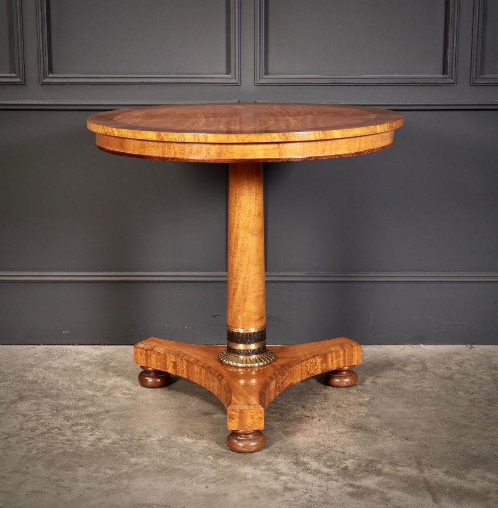 A circular antique satinwood occasional table featuring a crossbanded timber top, a central cylindrical pedestal with a brass mount, and a triform base on bun feet. View IV