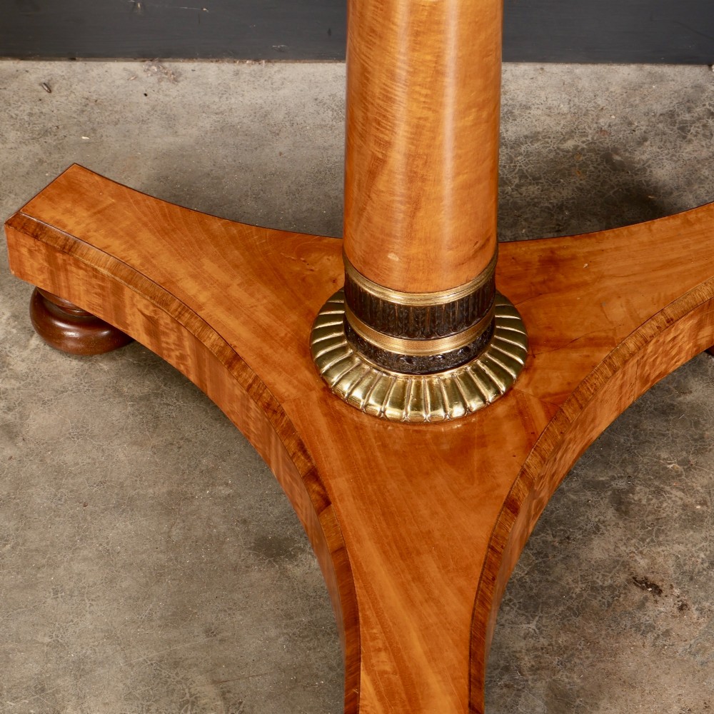 A circular antique satinwood occasional table featuring a crossbanded timber top, a central cylindrical pedestal with a brass mount, and a triform base on bun feet. View V