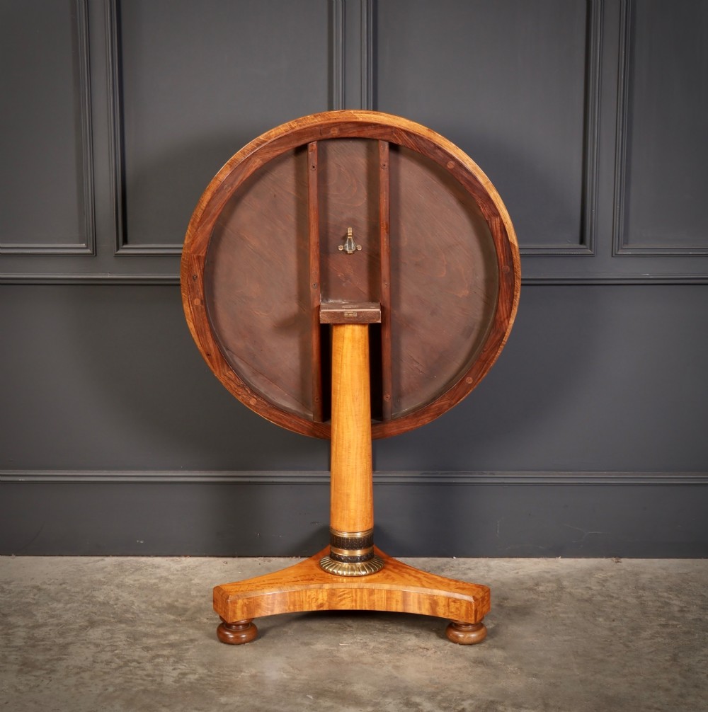 A circular antique satinwood occasional table featuring a crossbanded timber top, a central cylindrical pedestal with a brass mount, and a triform base on bun feet. View VII