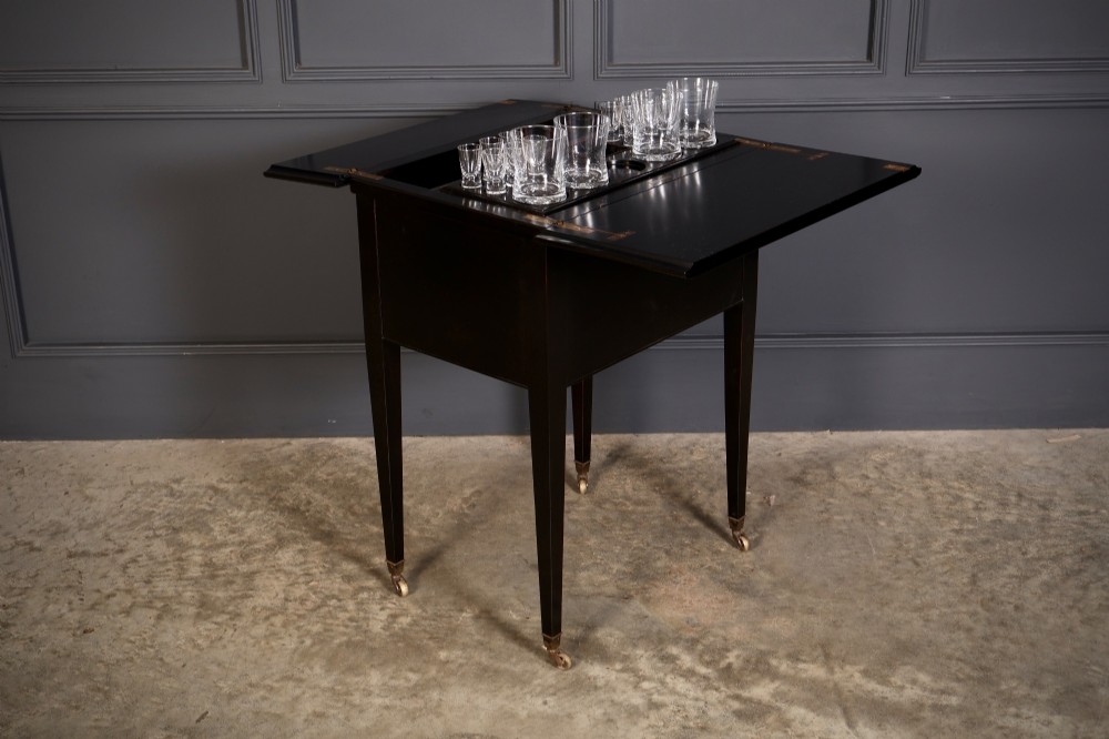 An ebonised wooden mechanical drinks table featuring opening top leaves that elevate a tray set with clear glassware, resting on tapered legs with brass castors.