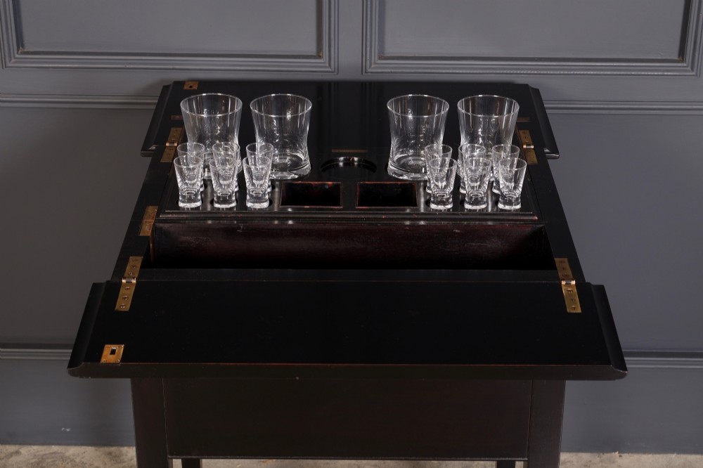 An ebonised wooden mechanical drinks table featuring opening top leaves that elevate a tray set with clear glassware, resting on tapered legs with brass castors. View III