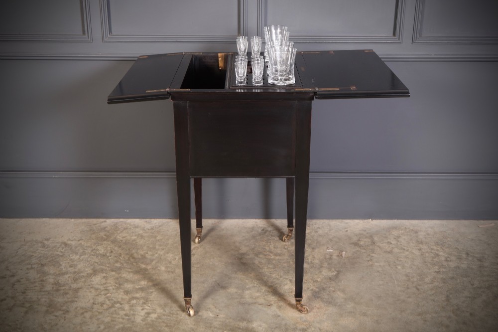 An ebonised wooden mechanical drinks table featuring opening top leaves that elevate a tray set with clear glassware, resting on tapered legs with brass castors. View V
