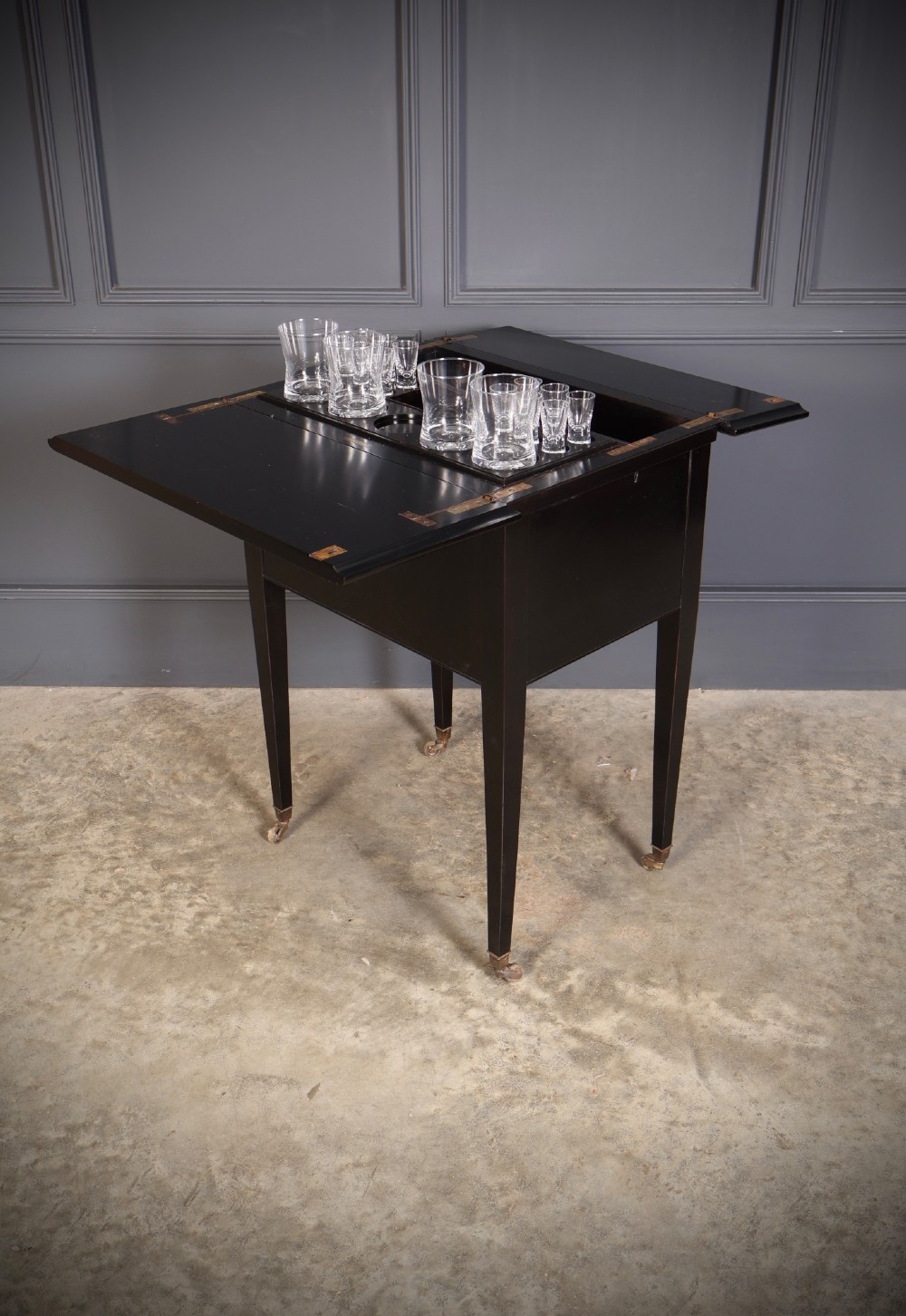 An ebonised wooden mechanical drinks table featuring opening top leaves that elevate a tray set with clear glassware, resting on tapered legs with brass castors. View VII