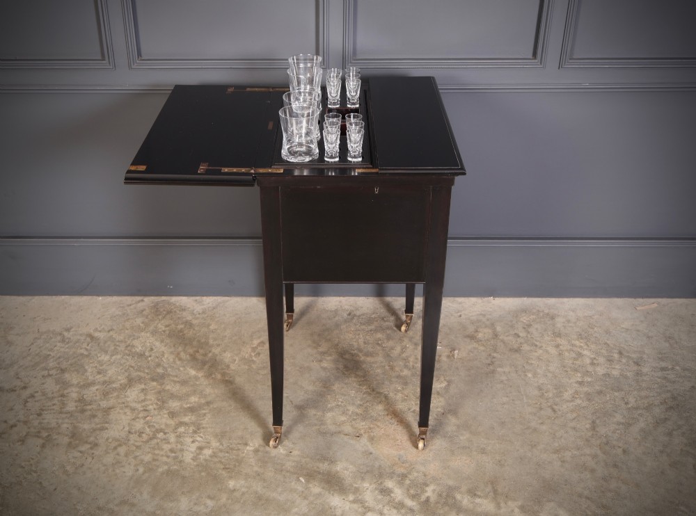 An ebonised wooden mechanical drinks table featuring opening top leaves that elevate a tray set with clear glassware, resting on tapered legs with brass castors. View VIII