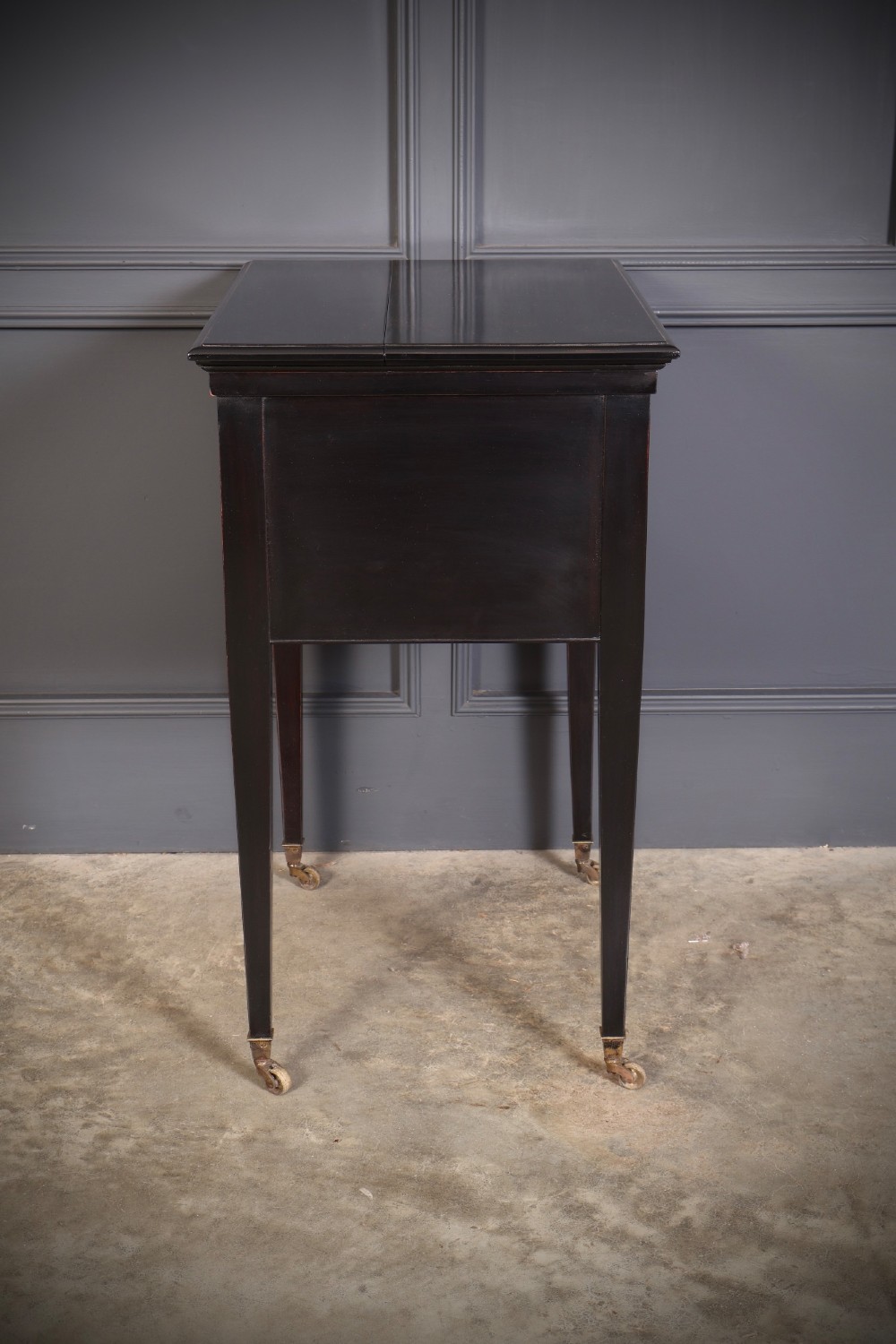 An ebonised wooden mechanical drinks table featuring opening top leaves that elevate a tray set with clear glassware, resting on tapered legs with brass castors. View XII