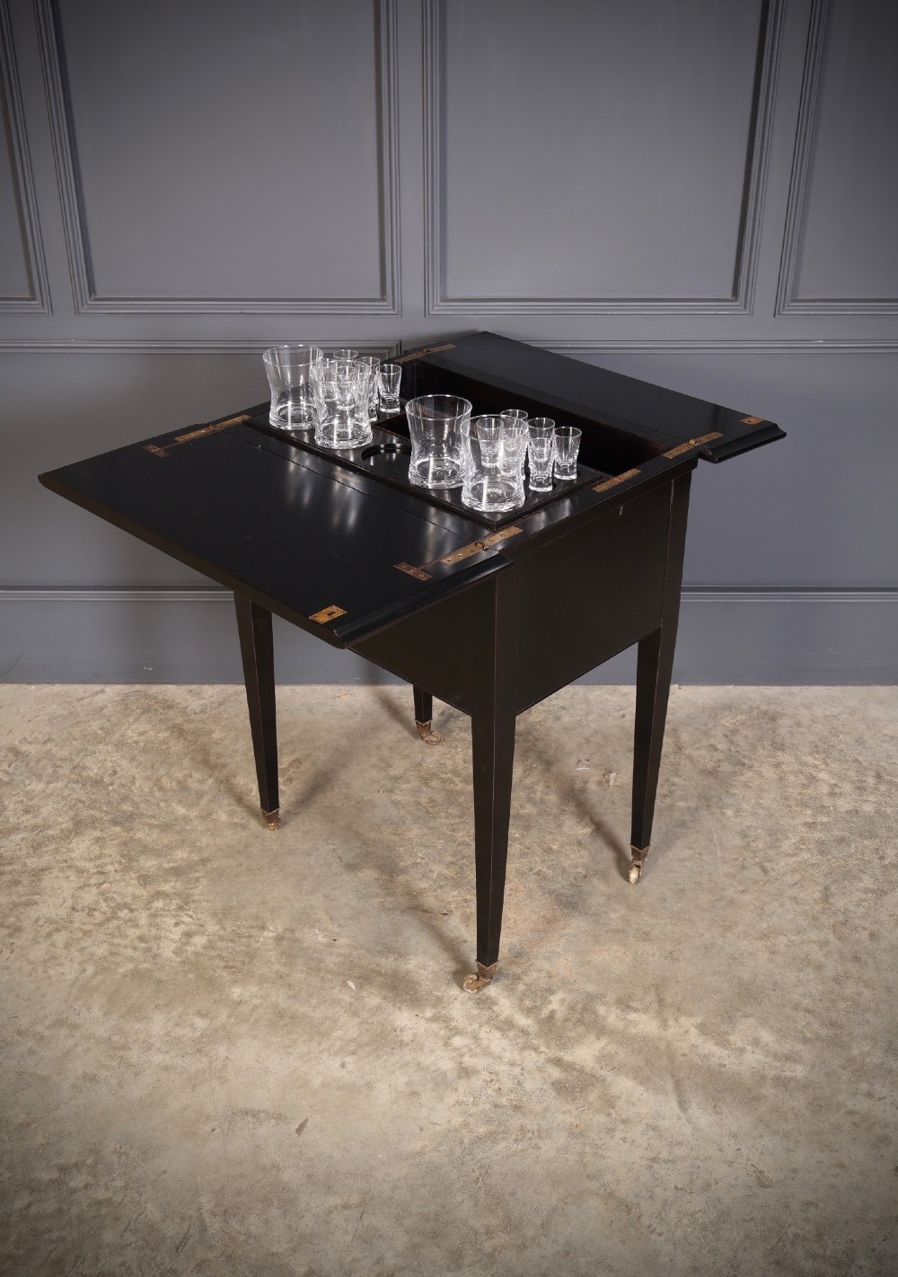 An ebonised wooden mechanical drinks table featuring opening top leaves that elevate a tray set with clear glassware, resting on tapered legs with brass castors. View XIV