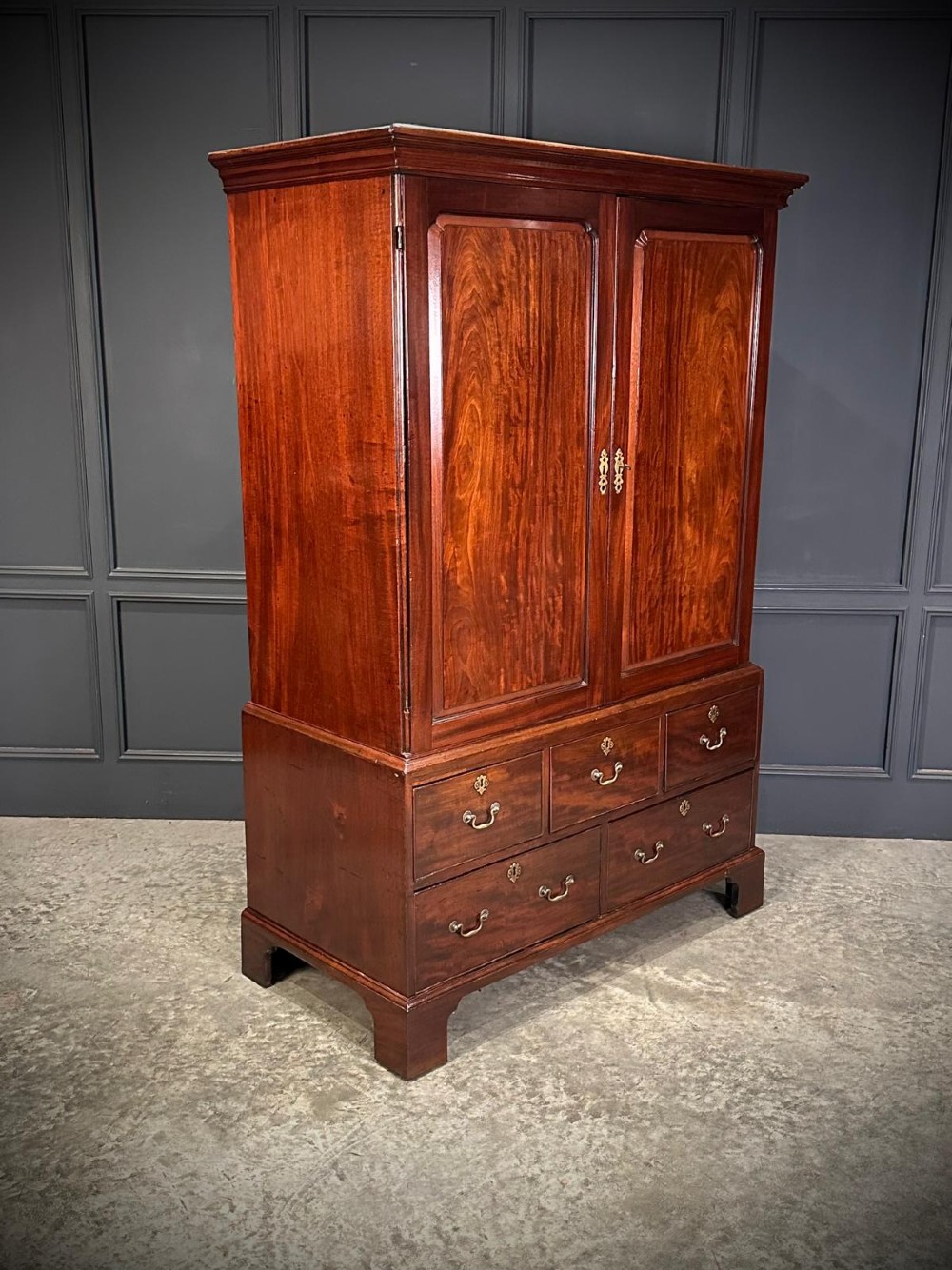 An antique Georgian mahogany gentlemans wardrobe featuring panelled upper cupboard doors above five lower drawers with brass swan-neck handles, resting on bracket feet. View II