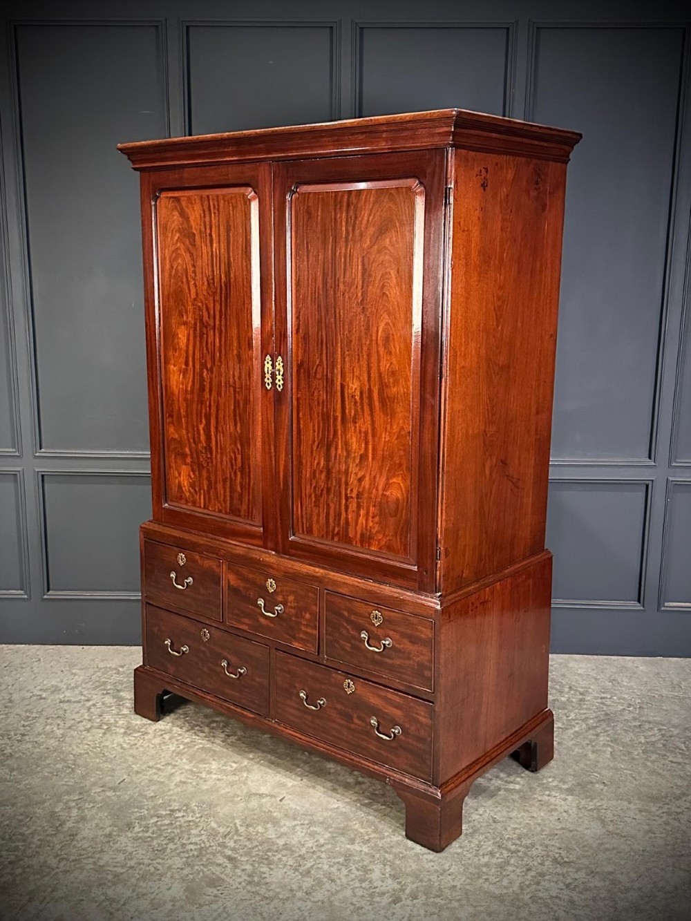 An antique Georgian mahogany gentlemans wardrobe featuring panelled upper cupboard doors above five lower drawers with brass swan-neck handles, resting on bracket feet.