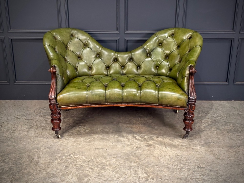 A Victorian double spoon-back settee upholstered in deep buttoned green leather with a carved mahogany frame set on brass castors. View II