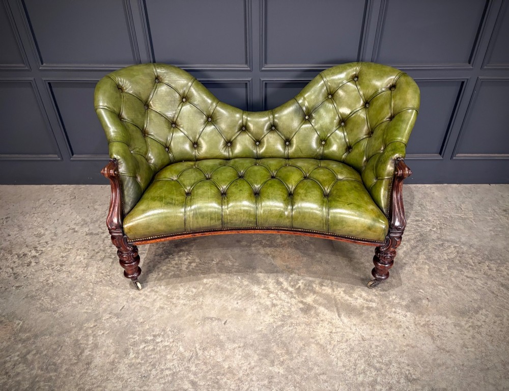 A Victorian double spoon-back settee upholstered in deep buttoned green leather with a carved mahogany frame set on brass castors. View VI